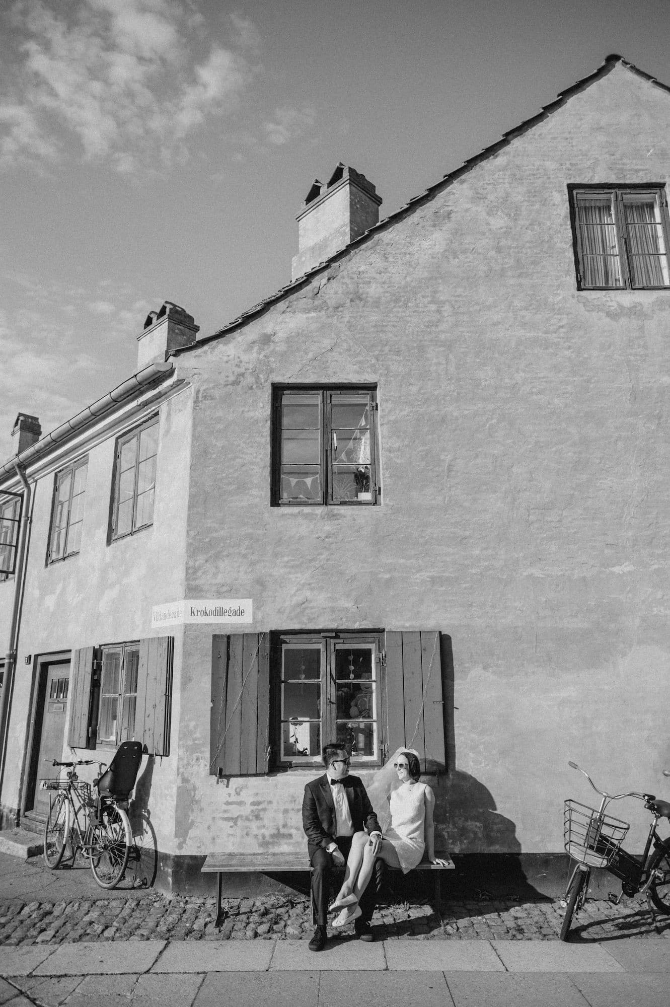A couple seated on a bench in front of a house with bicycles nearby in a Danish setting.