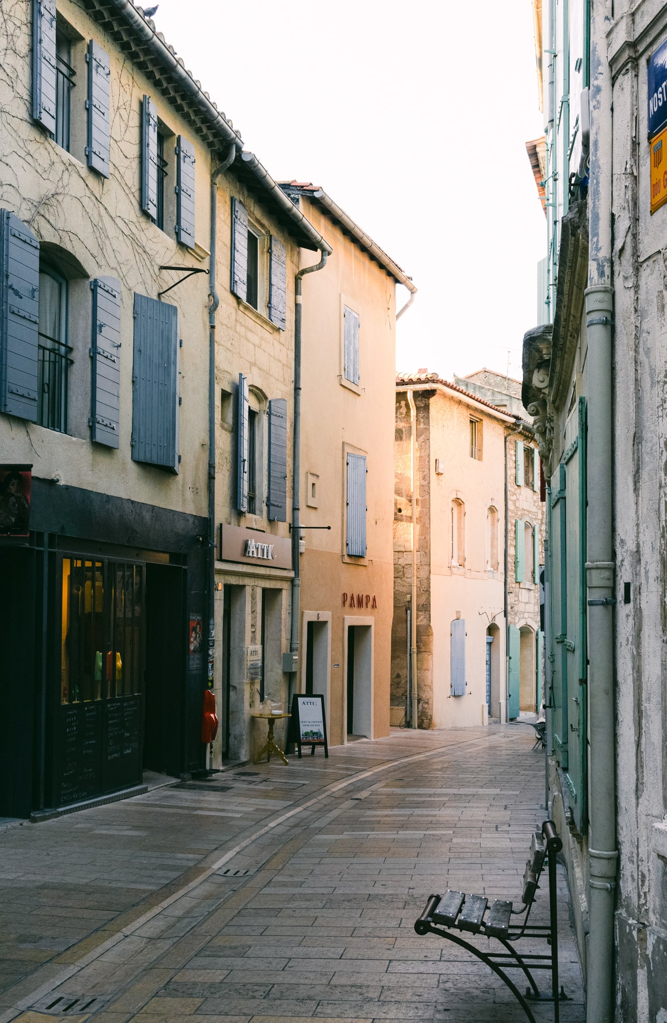 Narrow street lined with buildings featuring shutters and a bench in a Southern French town.