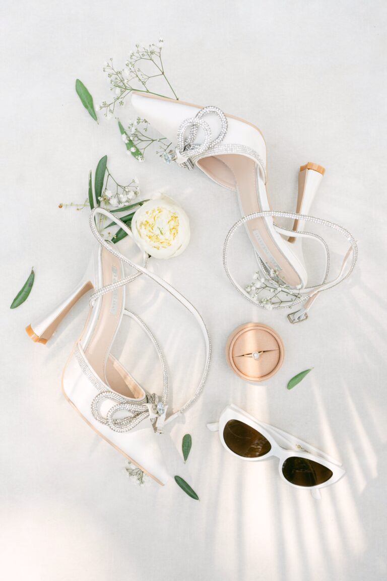 White high-heeled shoes adorned with decorative accents, surrounded by small flowers and sunglasses.