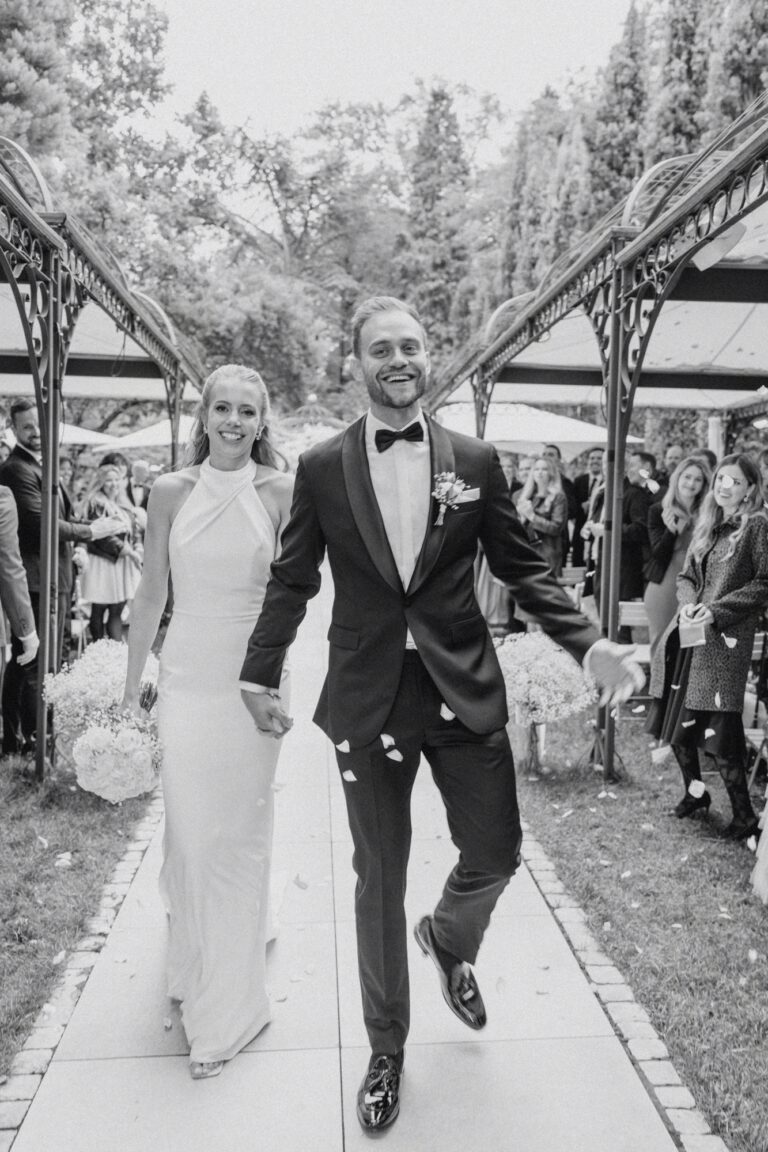 Bride in elegant dress and groom in tuxedo walking hand in hand after wedding ceremony with guests in the background.
